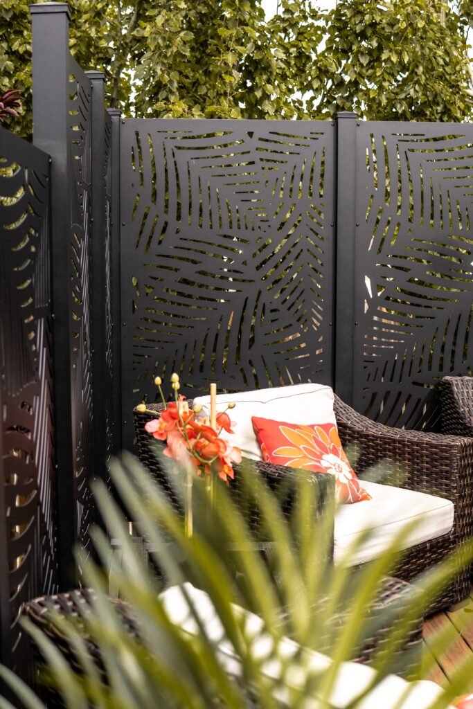 Stilvolle Außenecke mit Rattansessel, weißem Kissen und rotem Dekokissen. Ein künstlerisch durchbrochener, schwarzer Metallzaun umgibt den Bereich, umgeben von üppigem Grün und Palmenblättern, die tropisches Flair vermitteln.