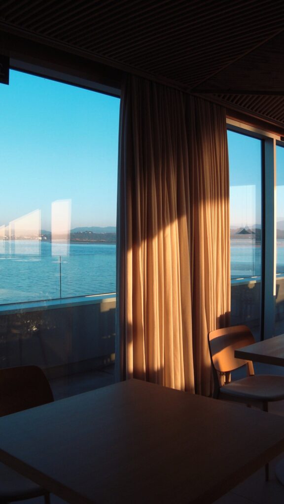 Gemütlicher Raum mit Fensterblick auf eine Bucht bei Sonnenuntergang, abgeschirmt durch lange, beige Vorhänge.