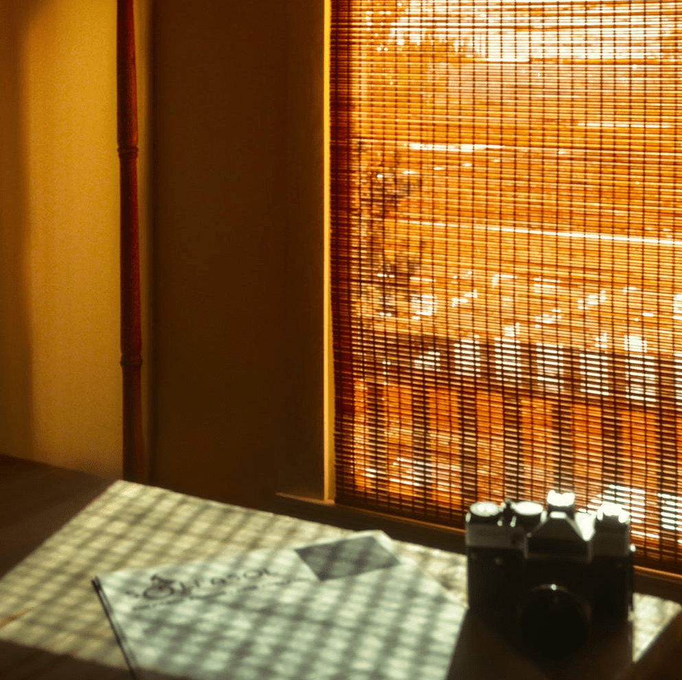 Vintage schwarze Kamera auf einem Tisch mit gemustertem Tischtuch, beleuchtet durch Sonnenlicht, das durch ein Bambusrollo in einem gemütlichen Raum filtert.