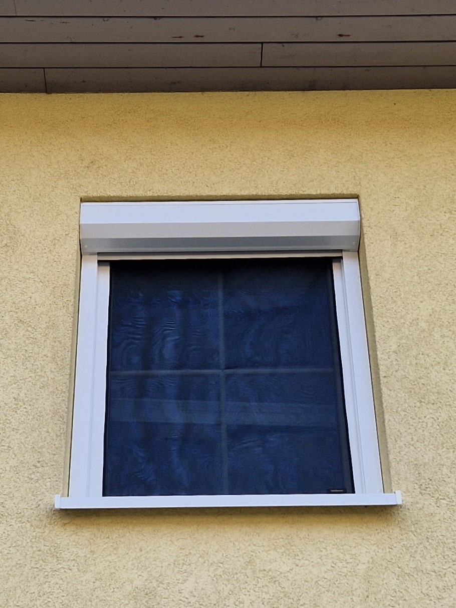 Fenster mit weißem Rahmen und außenliegendem schwarzem Gewebe-Hitzeschutz in gelber Hausfassade – Außenansicht